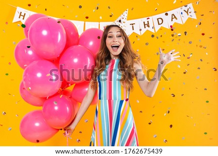 Image of excited cute woman in party cone posing with pink balloons isolated over yellow background