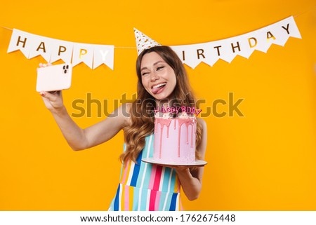 Image of amusing woman showing torte and taking selfie on cellphone isolated over yellow background