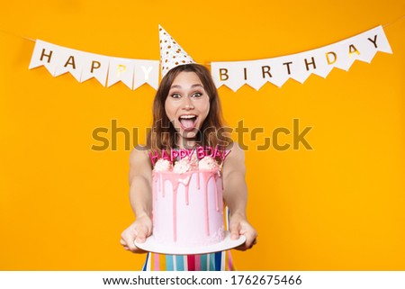 Image of excited young woman in party cone showing birthday torte and smiling isolated over yellow background