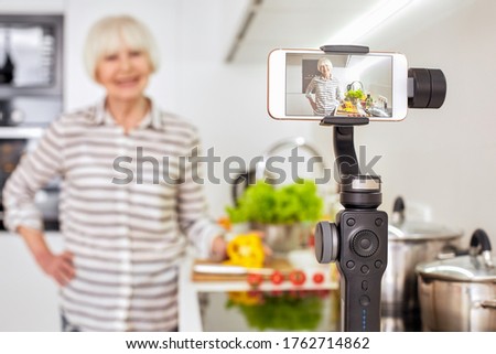 Atractive 70s years old woman blogger cook recording video kitchen with food smile to camera. Blurred background. White beauty senior teach show recipe look make photo stories for vlog Hobby broadcast