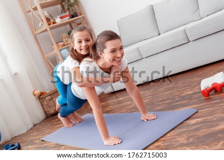 Daughter fools in funny pose while lying on mother's back. Looks into camera in studio background. Study teach new fitness sports exercises, lying on floor in middle of home interior room. Horizontal.