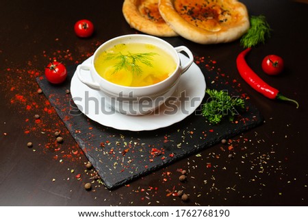   Overhead photo of a plate of chicken soup taken from above on a dark rustic texture with a spoon, a wooden ladle with pepper, slices of bread, and a place for text.
