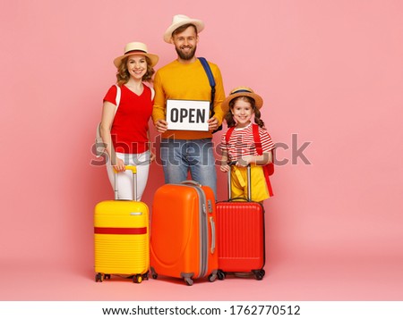 Full body of delighted parents and daughter with suitcases and Open sign smiling and looking at camera during summer vacation against pink backdrop

