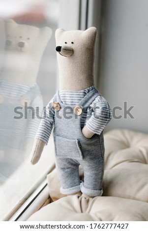 Cute childish toy Scandinavian bear stands at the window in overalls gray color 