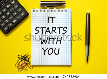 White note with inscription 'it starts with you' on beautiful yellow background, colored paper clips, metalic pen, cap and calculator. Business concept.