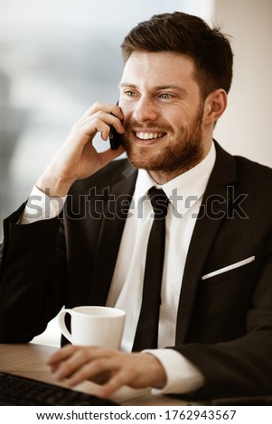 Business concept. Young businessman sitting at the office table happy talking on a cell phone getting good news about his work. Man in suit indoors on glass window background.