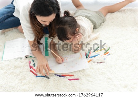 top view of happiness Asian mother teaching her little daughter to draw by pencil and crayon, lie on the floor of the house, family joyful together in the house on holiday
