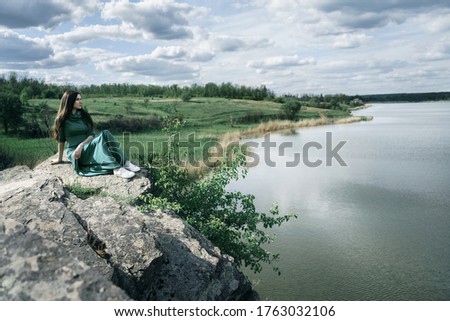 Girl in green dress sits on a rock above the water and looking from away. Russian landscape