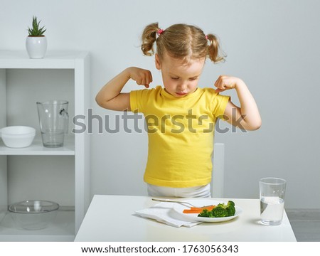 skeptic child weakly believes what it will be strong after eating broccoli. Super power broccoli, healthy baby, yellow t-shirt, white room, plate of vegetables. Royalty-Free Stock Photo #1763056343