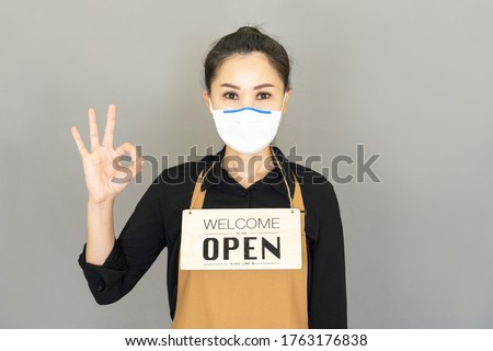 Young Small business owner wore medical face mask,brown apron and  the sign for the reopening of the place after the quarantine,Show her hand as OK symbols.