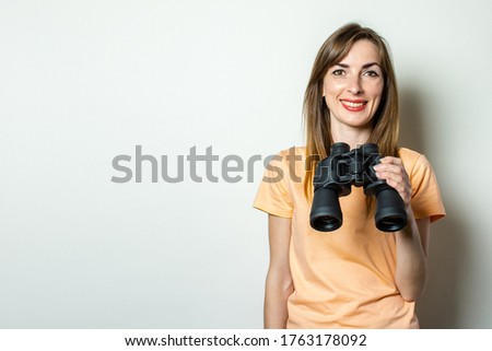 Young friendly girl in a T-shirt holds binoculars on a light background
