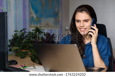 Overjoyed young woman in denim shirt working home with a laptop, talking via phone and looks to the laptop. Blur world map in the background