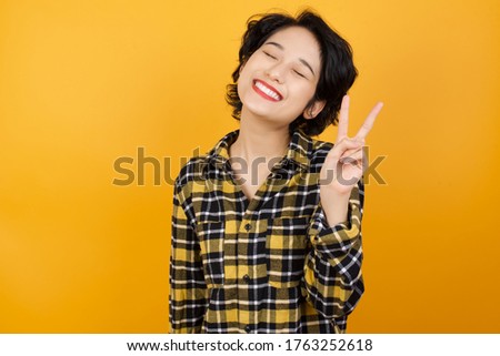 Young asian woman with short hair wearing plaid shirt standing over yellow background smiling with happy face winking at the camera doing victory sign. Number two.