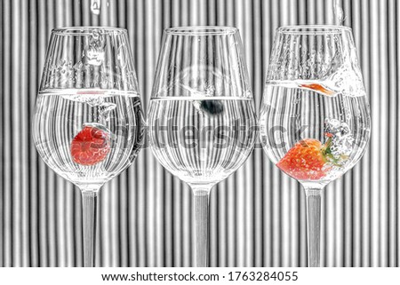 Three wine glasses half full with water, a raspberry, blueberry and strawberry being dropped into them, on a striped background. Artistic edit with intentional grain. Selective colour/black and white