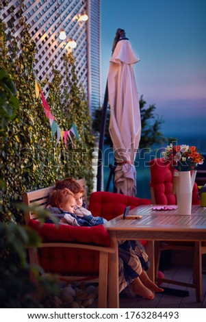cute siblings having fun, watching cartoons on the phone, spending time on cozy rooftop patio on summer evening