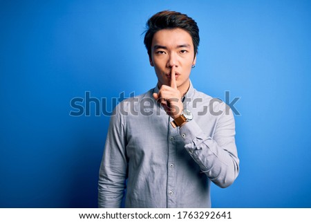 Young handsome chinese man wearing casual shirt standing over isolated blue background asking to be quiet with finger on lips. Silence and secret concept.