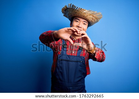 Young handsome chinese farmer man wearing apron and straw hat over blue background smiling in love doing heart symbol shape with hands. Romantic concept.