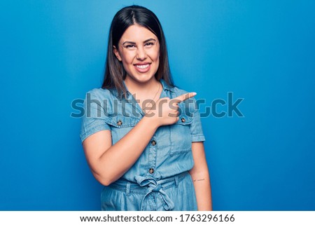 Young beautiful brunette woman wearing casual denim dress over isolated blue background smiling cheerful pointing with hand and finger up to the side