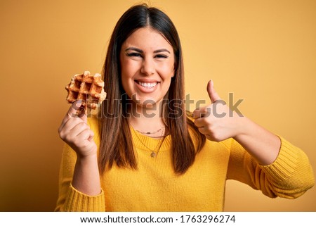 Young beautiful woman eating sweet waffle pastry over yellow background happy with big smile doing ok sign, thumb up with fingers, excellent sign