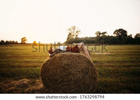 Stylish girl relaxing on hay bale in summer field in sunset. Young woman in hat resting on hay in sunshine, atmospheric tranquil moment. Countryside slow life Royalty-Free Stock Photo #1763298794