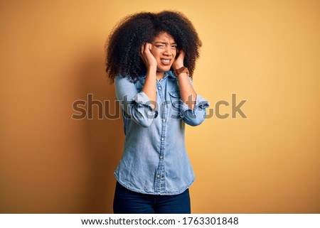 Young beautiful african american woman with afro hair standing over yellow isolated background covering ears with fingers with annoyed expression for the noise of loud music. Deaf concept.