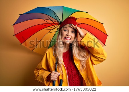 Young beautiful blonde woman wearing raincoat and wool cap holding colorful umbrella stressed with hand on head, shocked with shame and surprise face, angry and frustrated. Fear and upset for mistake.