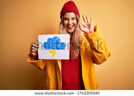 Young beautiful blonde woman wearing raincoat holding banner with cloud thunder image doing ok sign with fingers, excellent symbol