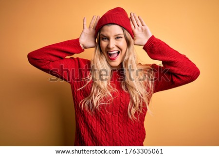 Young beautiful blonde woman wearing casual sweater and wool cap over white background Smiling cheerful playing peek a boo with hands showing face. Surprised and exited