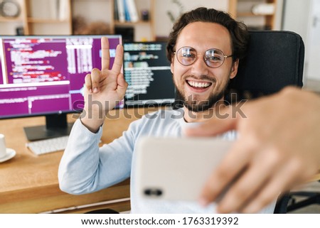 Image of unshaven joyful programmer man gesturing peace sign while taking selfie photo on cellphone in office