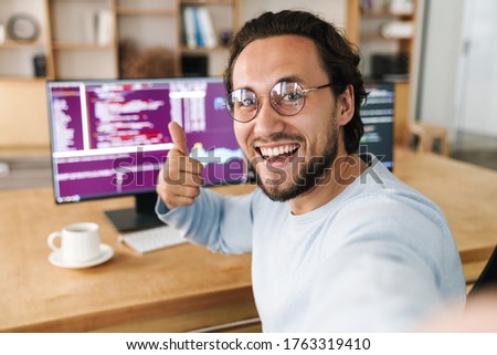 Image of unshaven excited programmer man showing thumb up while taking selfie photo in office