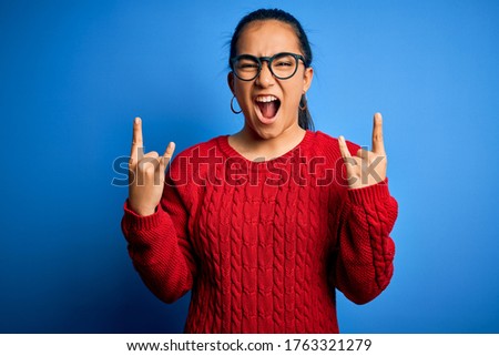 Young beautiful asian woman wearing casual sweater and glasses over blue background shouting with crazy expression doing rock symbol with hands up. Music star. Heavy concept.