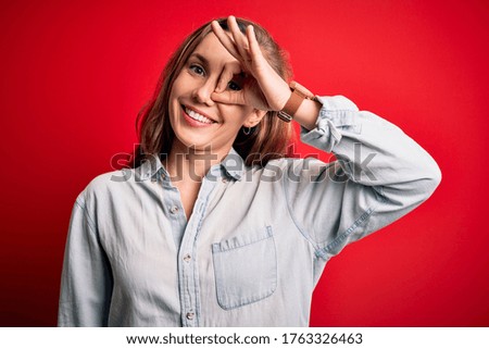 Young beautiful blonde woman wearing casual denim shirt over isolated red background doing ok gesture with hand smiling, eye looking through fingers with happy face.