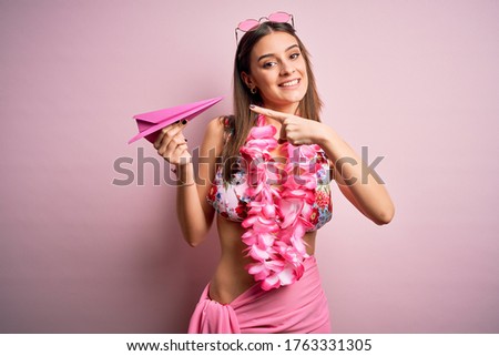 Young beautiful woman on vacation wearing bikini and hawaiian lei holding paper airplane very happy pointing with hand and finger