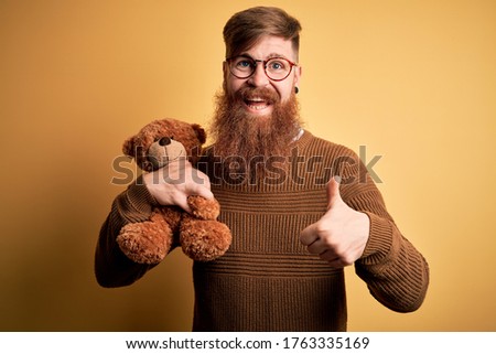 Redhead Irish man with beard hugging teddy bear stuffed animal over yellow isolated background happy with big smile doing ok sign, thumb up with fingers, excellent sign