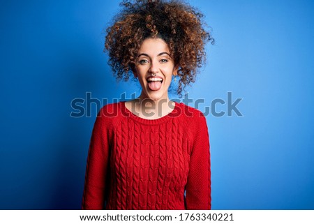 Young beautiful woman with curly hair and piercing wearing casual red sweater sticking tongue out happy with funny expression. Emotion concept.