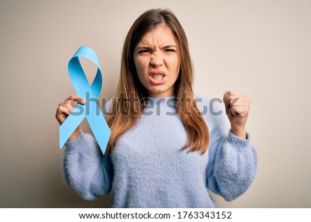 Young blonde woman holding prostate cancer awareness campaing blue ribbon annoyed and frustrated shouting with anger, crazy and yelling with raised hand, anger concept