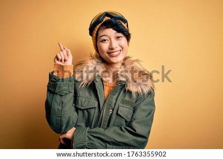 Beautiful asian skier girl wearing snow sportswear using ski goggles over yellow background with a big smile on face, pointing with hand and finger to the side looking at the camera.