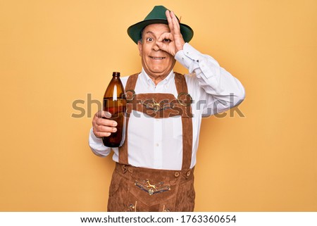 Senior grey-haired man wearing german traditional octoberfest suit drinking bottle of beer with happy face smiling doing ok sign with hand on eye looking through fingers