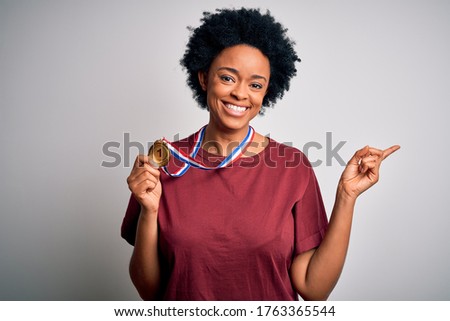 Young African American athlete woman with curly hair wearing gold medal winner competition very happy pointing with hand and finger to the side
