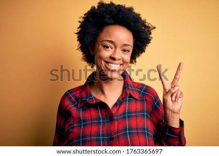 Young beautiful African American afro woman with curly hair wearing casual shirt smiling with happy face winking at the camera doing victory sign. Number two.