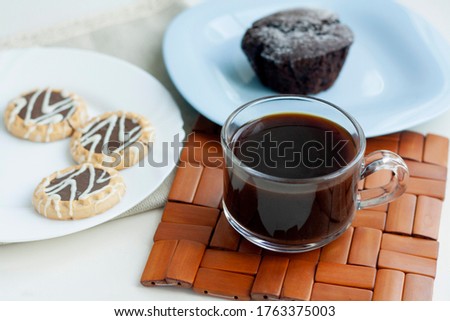 Breakfast, coffee in a glass cup on a wicker stand, chocolate muffin on a blue plate, cookies on a white plate, napkin