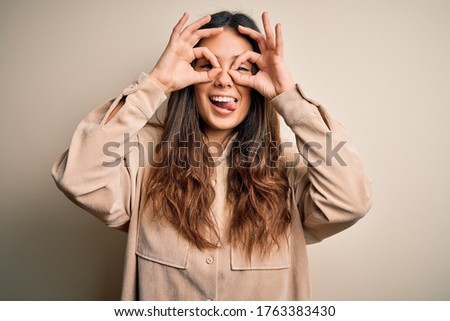 Young beautiful brunette woman wearing casual shirt standing over white background doing ok gesture like binoculars sticking tongue out, eyes looking through fingers. Crazy expression.