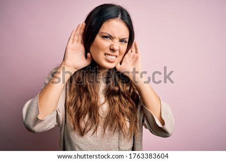 Young beautiful brunette woman wearing casual sweater standing over pink background Trying to hear both hands on ear gesture, curious for gossip. Hearing problem, deaf