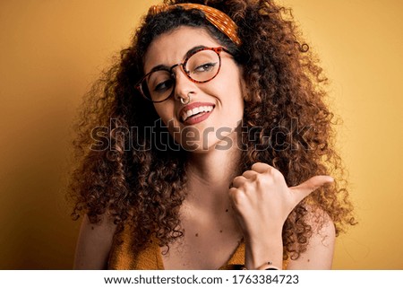 Beautiful brunette woman on vacation with curly hair and piercing wearing glasses and diadem smiling with happy face looking and pointing to the side with thumb up.
