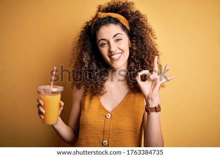 Young beautiful woman with curly hair and piercing drinking healthy orange juice doing ok sign with fingers, excellent symbol