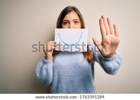 Young blonde woman holding funny smile drawing on mouth as happy expression with open hand doing stop sign with serious and confident expression, defense gesture
