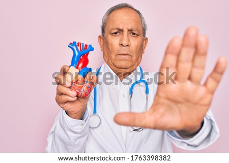 Senior hoary doctor man wearing stethoscope holding plastic heart over pink background with open hand doing stop sign with serious and confident expression, defense gesture