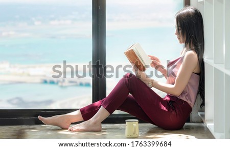 Young beautiful asian woman reading a book alone while sitting on the floor near the window in living room. Copy space.