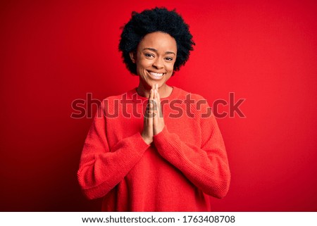 Young beautiful African American afro woman with curly hair wearing casual sweater praying with hands together asking for forgiveness smiling confident.