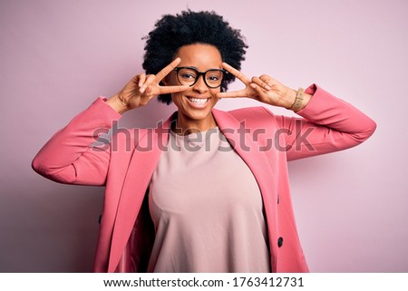 Young beautiful African American afro businesswoman with curly hair wearing pink jacket Doing peace symbol with fingers over face, smiling cheerful showing victory
